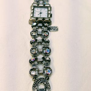 May include: A silver-toned analog watch with a square white face featuring black numerals at 12, 3, 6, and 9. The band consists of interconnected circular links embedded with iridescent clear crystals, finished with a crescent moon charm.