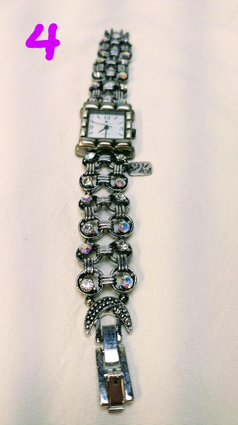 May include: A silver-toned analog watch with a square white face featuring black numerals at 12, 3, 6, and 9. The band consists of interconnected circular links embedded with iridescent clear crystals, finished with a crescent moon charm.