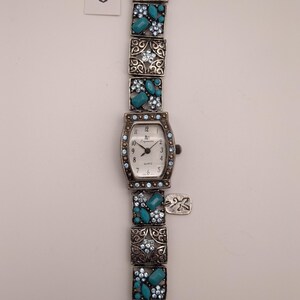 May include: A silver-tone quartz watch with a rectangular face featuring black numerals and the text "Espritime". The band consists of square links decorated with light blue rhinestones and turquoise-colored stones, with a small metal tag attached.