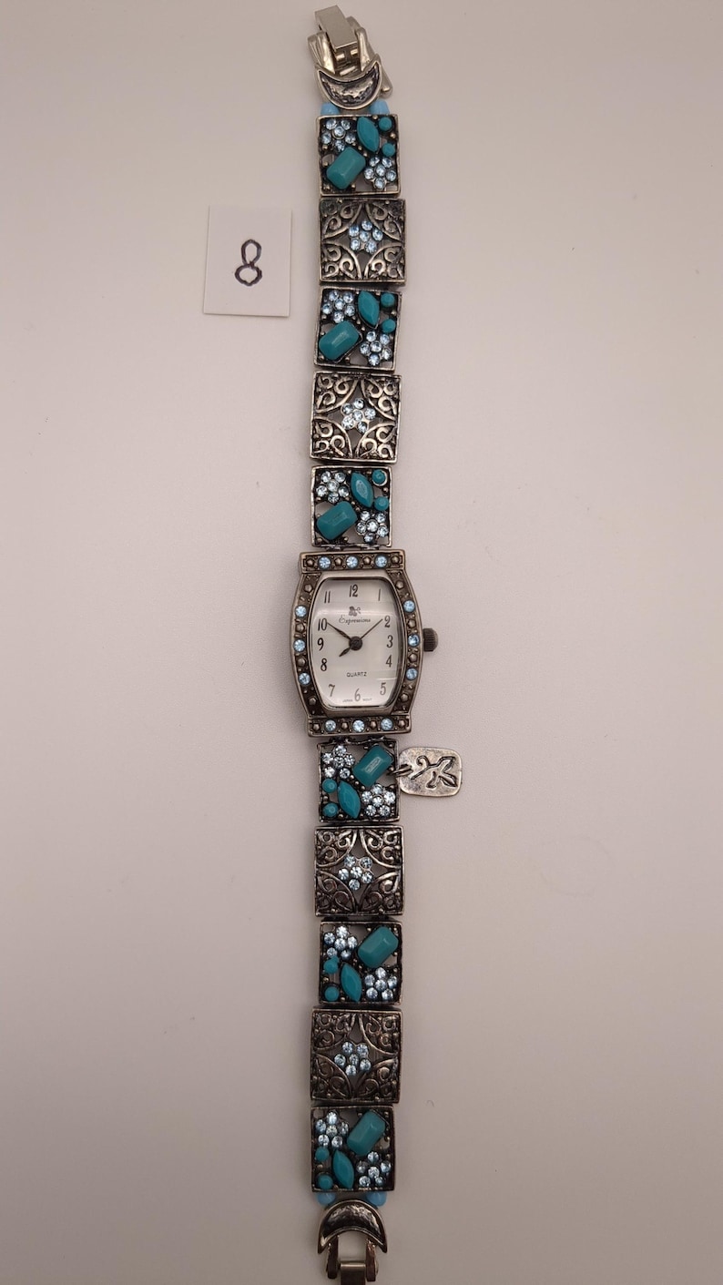 May include: A silver-tone quartz watch with a rectangular face featuring black numerals and the text "Espritime". The band consists of square links decorated with light blue rhinestones and turquoise-colored stones, with a small metal tag attached.