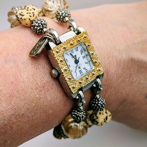May include: A rectangular wristwatch with a gold-tone engraved bezel and a white face featuring black Roman numerals. The watch band consists of two strands of beads, including tan speckled heart-shaped beads and translucent ribbed beads.