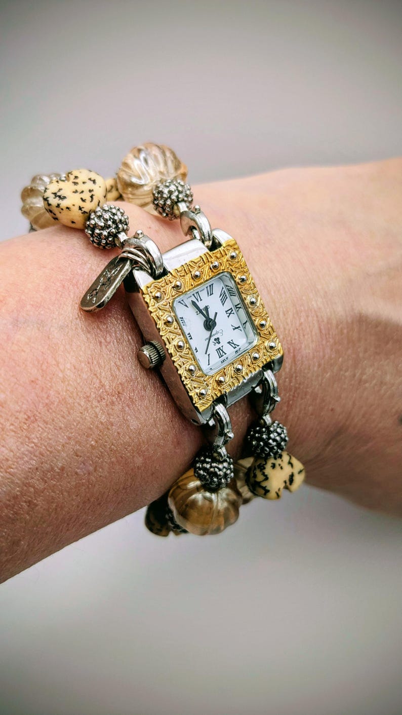 May include: A rectangular wristwatch with a gold-tone engraved bezel and a white face featuring black Roman numerals. The watch band consists of two strands of beads, including tan speckled heart-shaped beads and translucent ribbed beads.