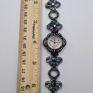 May include: A silver-toned analog wristwatch with a link bracelet featuring circular frames and bow-shaped accents embedded with purple crystals. The watch face is round with a light background. A wooden ruler shows the watch is 7 inches long.