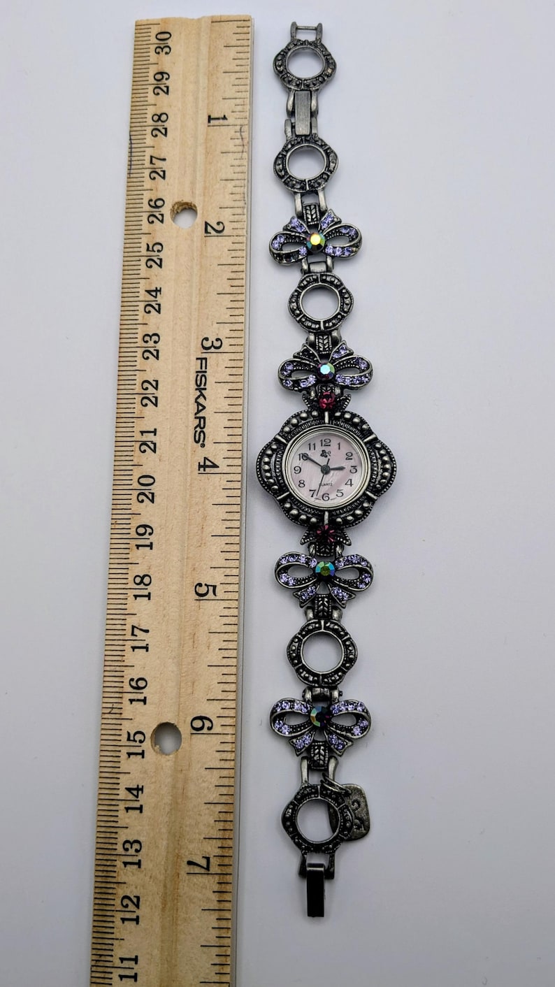 May include: A silver-toned analog wristwatch with a link bracelet featuring circular frames and bow-shaped accents embedded with purple crystals. The watch face is round with a light background. A wooden ruler shows the watch is 7 inches long.
