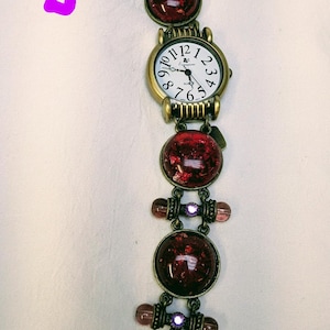 May include: A bronze-toned watch with a white face and black numerals, featuring a bracelet composed of round, dark red, glittery cabochons, small purple beads, and metal links. The watch includes a toggle clasp closure at the end of the band.