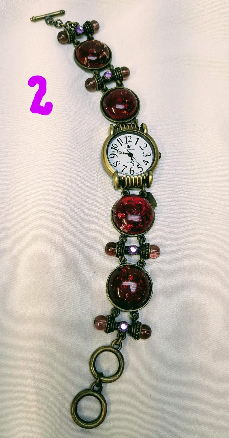 May include: A bronze-toned watch with a white face and black numerals, featuring a bracelet composed of round, dark red, glittery cabochons, small purple beads, and metal links. The watch includes a toggle clasp closure at the end of the band.