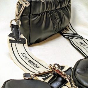 May include: Black crossbody bag with a detachable strap featuring the brand name "STEVE MADDEN" in white. The bag has a quilted design and a small, round, zippered pouch attached. The bag has a top handle and gold-colored hardware.