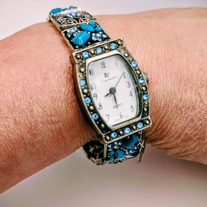 May include: A quartz wristwatch with a rectangular white face featuring black numerals and the brand name Expressions. The metal frame and bracelet are decorated with small light blue crystals and larger turquoise-colored stones in a dark setting.