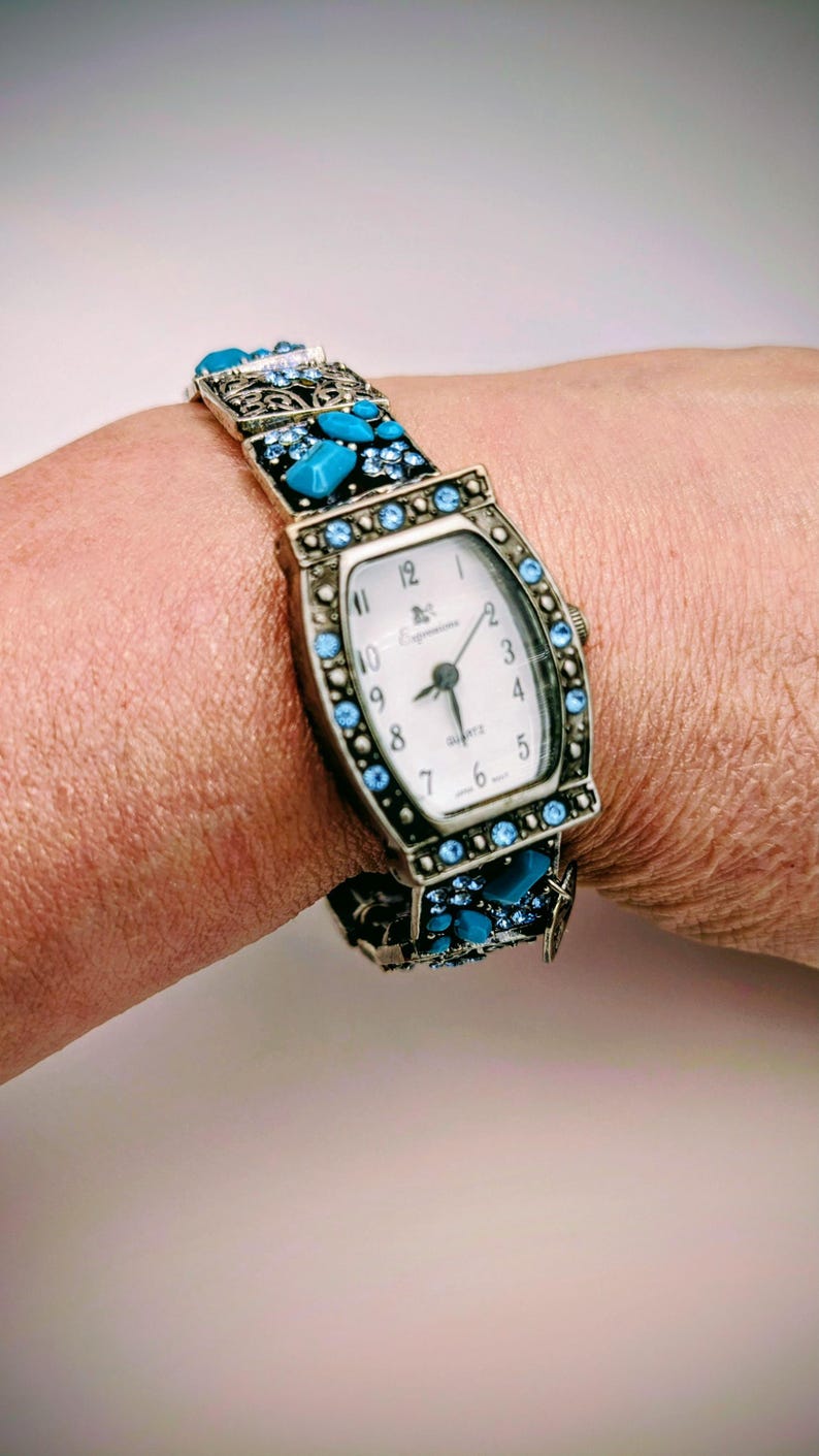 May include: A quartz wristwatch with a rectangular white face featuring black numerals and the brand name Expressions. The metal frame and bracelet are decorated with small light blue crystals and larger turquoise-colored stones in a dark setting.