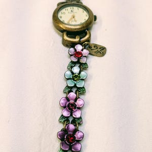 May include: A bronze-toned analog wristwatch featuring a round face with a white dial and gold hands. The band is composed of a series of connected metal flower charms in shades of purple, light blue, and pink, accented with small embedded crystals.