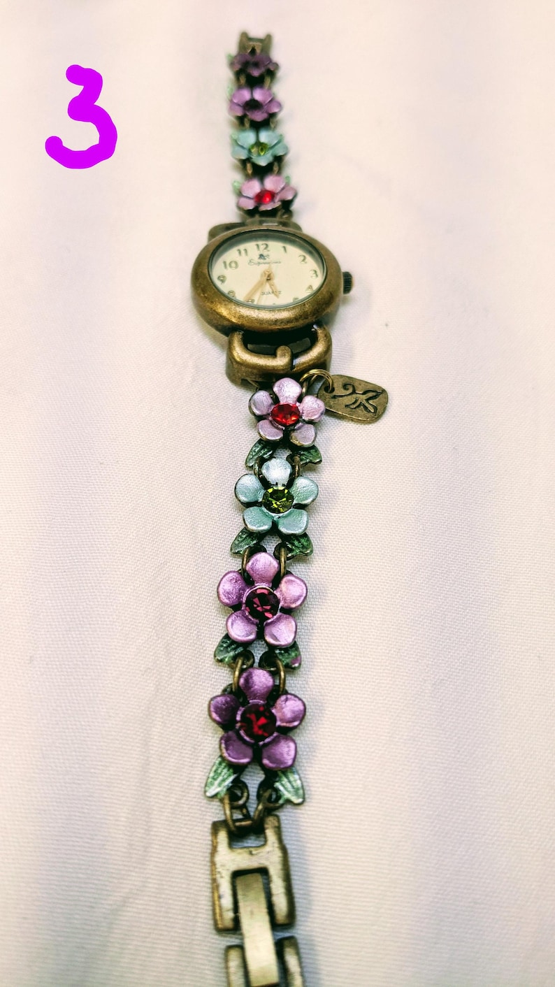 May include: A bronze-toned analog wristwatch featuring a round face with a white dial and gold hands. The band is composed of a series of connected metal flower charms in shades of purple, light blue, and pink, accented with small embedded crystals.