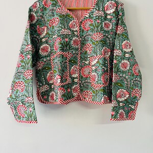 Può includere: Una giacca trapuntata con base azzurra e un motivo floreale ripetuto nei toni del rosa, rosso e verde. La giacca presenta maniche lunghe, scollo a V, due tasche frontali applicate e bordi rifiniti con una striscia rossa e bianca.
