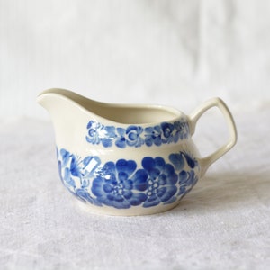 Op de afbeelding: Crèmekleurige keramische creamer met een blauw bloemmotief. De creamer heeft een tuit en een handvat. Het blauwe bloemmotief loopt rond de body van de creamer. De creamer staat op een wit oppervlak.