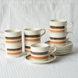 Può includere: Un set di tazze e piattini da caffè in ceramica. Le tazze e i piattini sono bianco sporco con bande orizzontali marroni e arancioni. Il set include tazze e piattini impilati, perfetti per servire caffè o tè. Ogni tazza contiene circa 240 ml.