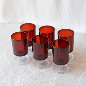 Può includere: Set di sei bicchieri da liquore in stile vintage. Ogni bicchiere presenta un interno rosso scuro e una base trasparente e arrotondata. I bicchieri sono cilindrici con un bordo leggermente svasato, perfetti per servire piccole porzioni.