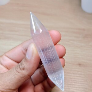 May include: A clear selenite crystal wand, held in a hand. The crystal is translucent with visible internal striations and a pointed tip. The background is a light-coloured wooden surface.