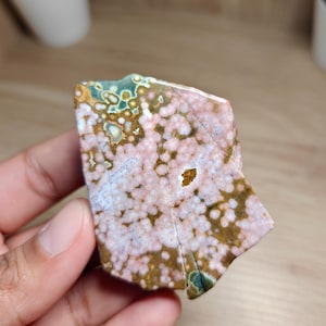 May include: A polished gemstone with a distinctive pattern. The stone displays a mix of colours, including pink, brown, green, and white. The surface is speckled with small, round patterns. The gemstone is held in a hand.