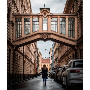 K&ouml;nnte beinhalten: Eine Frau geht eine enge Stra&szlig;e in einer europ&auml;ischen Stadt entlang. Die Stra&szlig;e ist von hohen Geb&auml;uden ges&auml;umt, und &uuml;ber der Stra&szlig;e befindet sich eine Br&uuml;cke. Die Br&uuml;cke ist aus Stein und hat einen dekorativen Torbogen. Die Frau tr&auml;gt einen langen Mantel und tr&auml;gt eine Tasche.