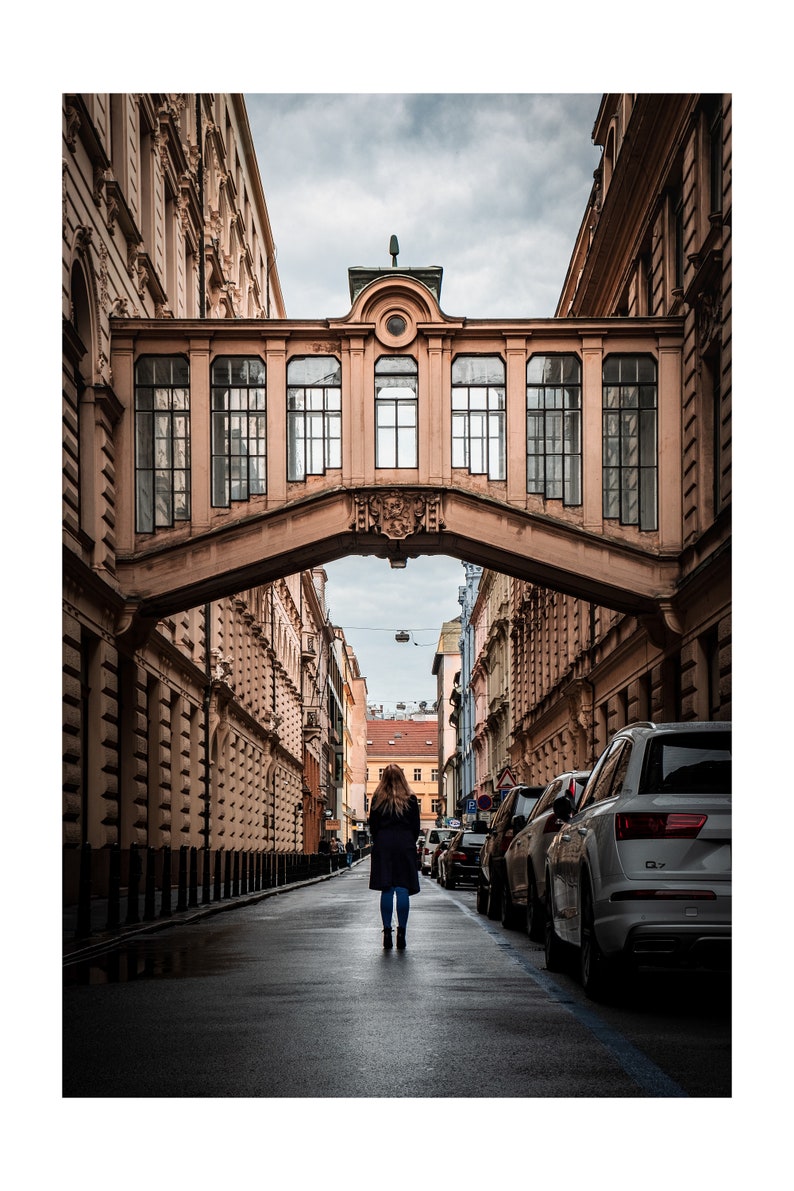 K&ouml;nnte beinhalten: Eine Frau geht eine enge Stra&szlig;e in einer europ&auml;ischen Stadt entlang. Die Stra&szlig;e ist von hohen Geb&auml;uden ges&auml;umt, und &uuml;ber der Stra&szlig;e befindet sich eine Br&uuml;cke. Die Br&uuml;cke ist aus Stein und hat einen dekorativen Torbogen. Die Frau tr&auml;gt einen langen Mantel und tr&auml;gt eine Tasche.