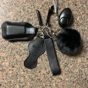 Può includere: Un portachiavi nero con una varietà di oggetti, tra cui una piccola bottiglia di disinfettante per le mani, un pon pon in pelliccia sintetica nero, un allarme di autodifesa nero e un portachiavi in ​​pelle nero.