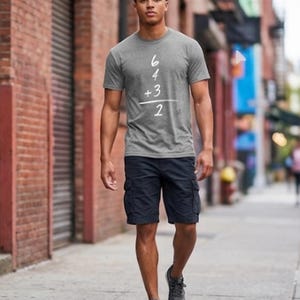 May include: A gray t-shirt with the numbers 6, 4, 3, and 2 arranged vertically with a plus sign and a line underneath. The person is wearing navy blue cargo shorts and gray sneakers. The person is walking on a sidewalk.