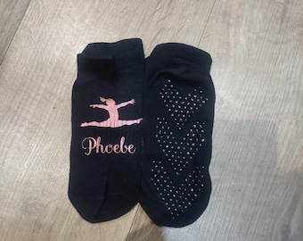 May include: Black ankle socks with a pink ballerina graphic and the name "Phoebe" printed on them. The soles have a pattern of small, silver non-slip grips. These socks are designed for dance or similar activities.