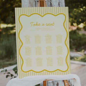 Puede incluir: Un diagrama de asientos a rayas amarillas y blancas con el texto "Take a seat" en un caballete de madera. El diagrama enumera los nombres y las asignaciones de mesa para una boda.