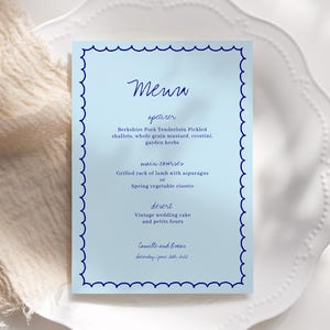 Könnte beinhalten: Eine blaue Speisekarte mit weißem Rand und einem gewellten Rand. Die Speisekarte trägt oben das Wort "Menu" in blauer Kursivschrift. Die Speisekarte listet Vorspeisen, Hauptgerichte und Dessert auf. Die Speisekarte ist für eine Hochzeit.