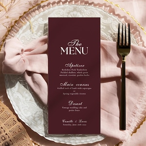 Può includere: Un biglietto del menu bordeaux con testo bianco, posto su un piatto bianco e dorato, con una forchetta dorata e un coltello nero. Il menu elenca antipasti, piatti principali e dessert. Sono visibili anche un tovagliolo rosa e un ventaglio decorativo.