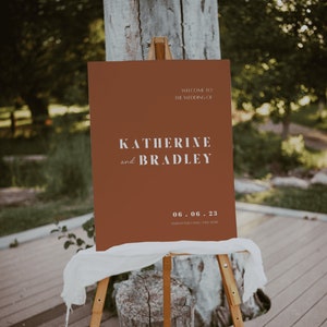 May include: A brown welcome sign with white text announcing the wedding of Katherine and Bradley on June 6, 2023. The sign is on a wooden easel.