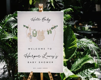 May include: White welcome sign with green foliage and a baby outfit illustration. The sign reads "Hello Baby", "Welcome to Harper Lewis's Baby Shower", and "09 June 2023".