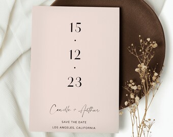 以下が含まれることがあります： 12.15.23の日付と、カミーユとアーサーの名前が筆記体で書かれた、薄いピンク色の結婚式の招待状。名前の下に「Save the Date」と「ロサンゼルス、カリフォルニア」の文字が印刷されている。