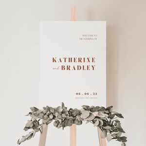May include: A white wedding sign with brown text announcing the wedding of Katherine and Bradley on June 6, 2023. The sign is propped up on a wooden easel with greenery in front of it.