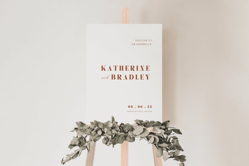 May include: A white wedding sign with brown text announcing the wedding of Katherine and Bradley on June 6, 2023. The sign is propped up on a wooden easel with greenery in front of it.
