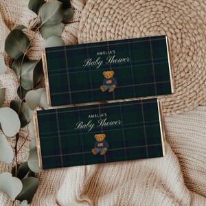 May include: Two chocolate bar wrappers with a green and navy plaid design. The wrappers feature the text "AMELIA'S Baby Shower" and a teddy bear graphic in a blue sweater and jeans. They are on a beige surface.