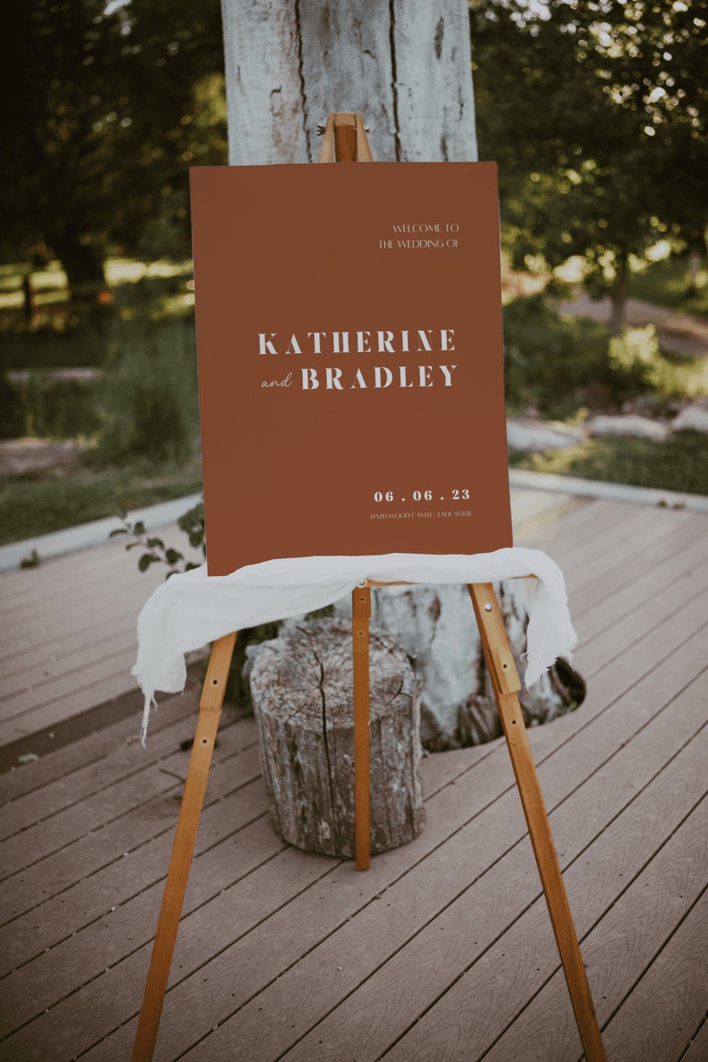 May include: A brown welcome sign with white text announcing the wedding of Katherine and Bradley on June 6, 2023. The sign is on a wooden easel.