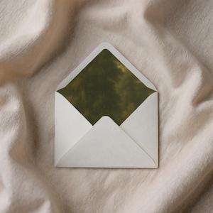 May include: A white envelope with a green tie-dye pattern on the inside. The envelope is open, revealing the patterned interior. The background is a beige, textured fabric.
