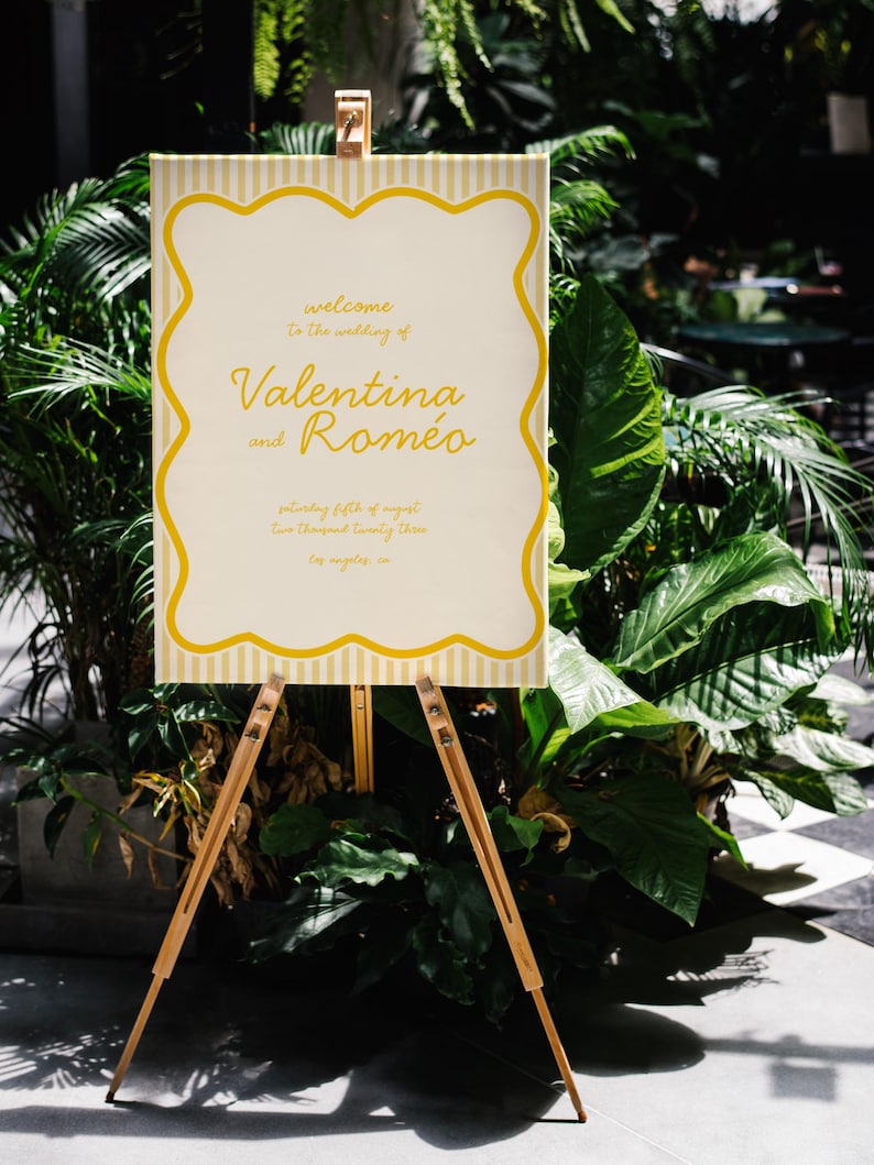 May include: A yellow and white striped sign with a yellow border and a white interior. The sign says "Welcome to the wedding of Valentina and Romeo, Saturday fifth of August, Two Thousand Twenty Three, Los Angeles, CA". The sign is on a wooden easel.