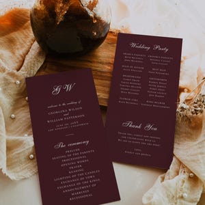 Può includere: Programmi di cerimonia nuziale bordeaux con testo bianco, con le iniziali della coppia e i dettagli del matrimonio. I programmi elencano gli invitati al matrimonio e l'ordine della cerimonia. Un vaso decorativo e tessuto sullo sfondo.