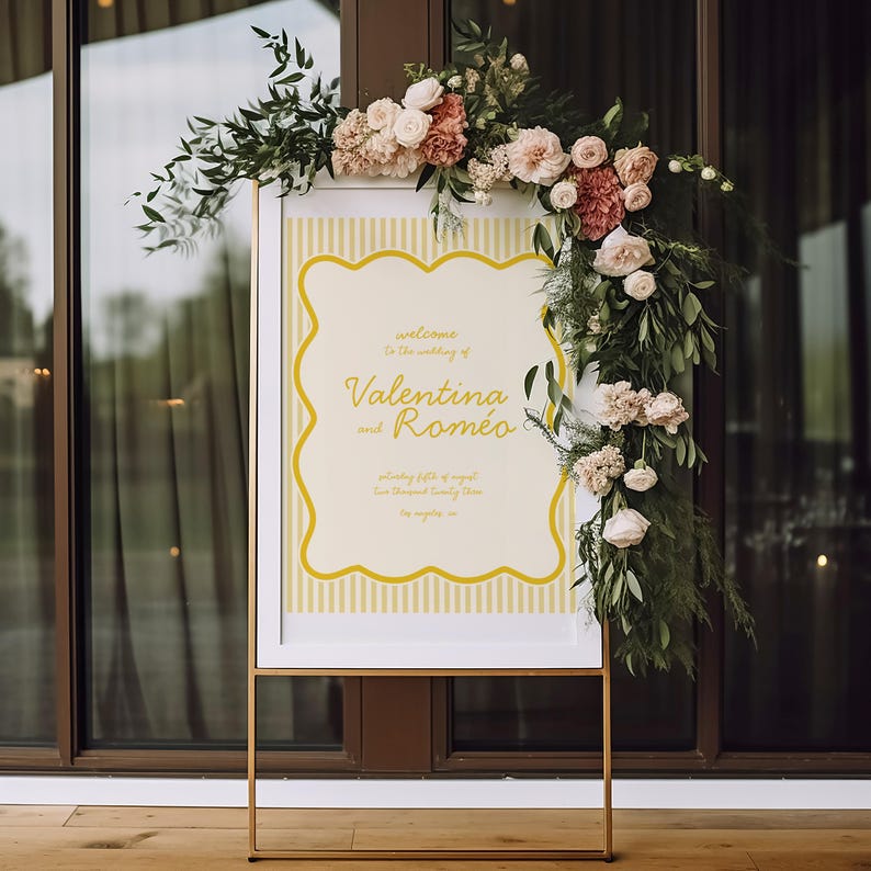 May include: A white framed sign with a yellow and white striped background. The sign has a gold border and says "welcome to the wedding of Valentina and Romeo" in gold lettering. The sign is decorated with a garland of pink and white flowers.