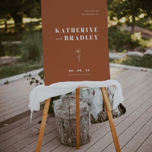 May include: A brown welcome sign with white text announcing the wedding of Katherine and Bradley on June 6, 2023 at Hawthorne Castle. A simple line drawing of a flower is featured on the sign.