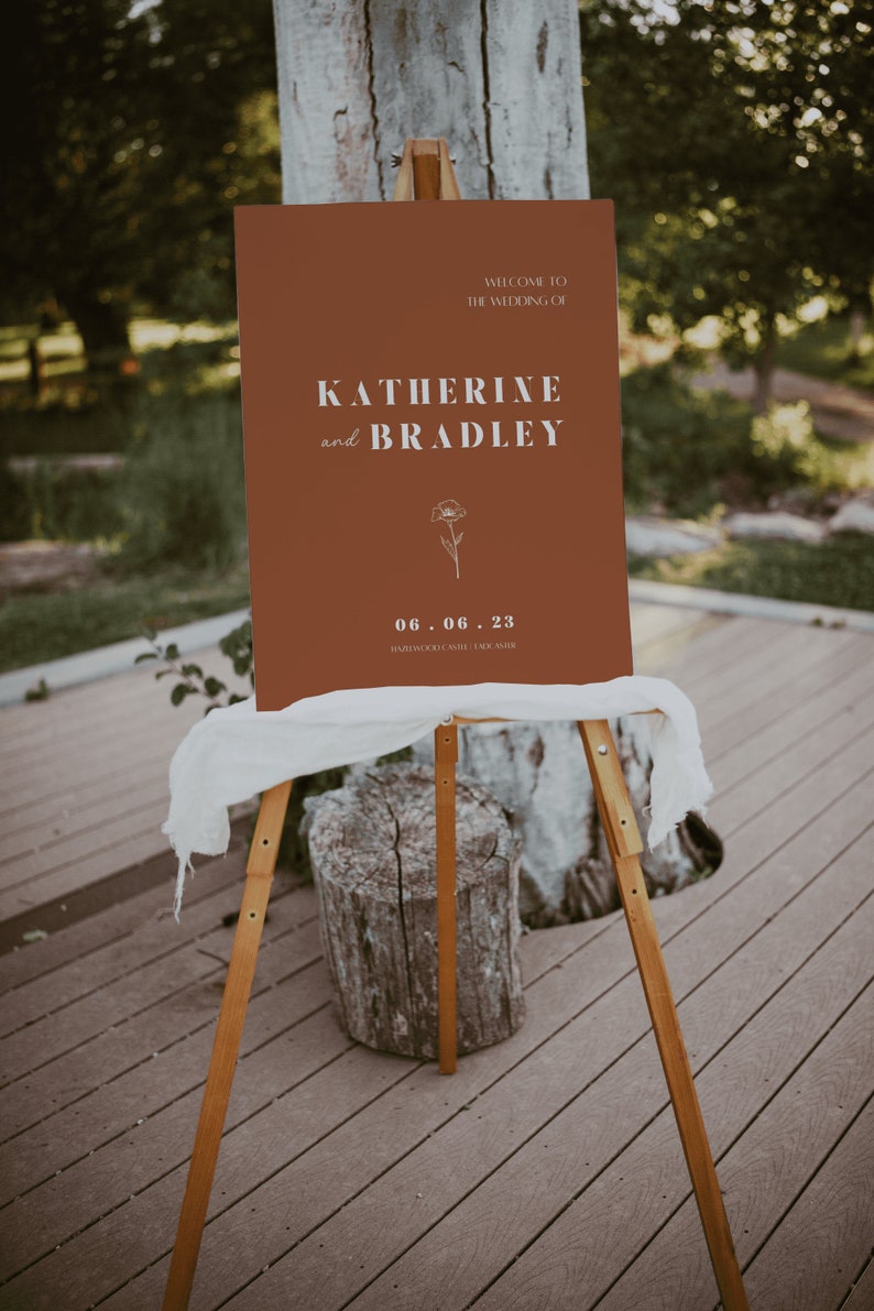 May include: A brown welcome sign with white text announcing the wedding of Katherine and Bradley on June 6, 2023 at Hawthorne Castle. A simple line drawing of a flower is featured on the sign.