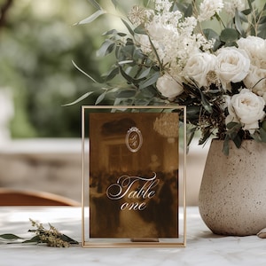 Puede incluir: Un letrero de mesa rectangular en un marco de metal dorado fino con una imagen en tono sepia marrón oscuro de una reunión formal. El texto "Table one" está impreso en letra cursiva blanca en el centro, con un pequeño emblema ovalado arriba.