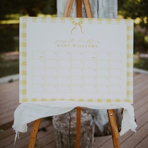 May include: A wooden easel displays a white sign with a yellow gingham border. The sign has a calendar grid and text that reads "guess the due date of BABY WILLIAMS." A white cloth drapes the easel.