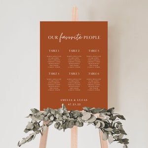 Peut inclure: Un tableau d'affectation des places de couleur rouille avec du texte blanc. Le tableau est intitulé "Nos personnes préférées" et liste les noms des invités assignés à chaque table. Le bas du tableau indique "Amelia & Lucas 07.23.22".