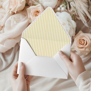 May include: A white envelope with a yellow and white striped interior is held open, revealing the pattern. The background features soft, light-colored flowers and fabric, creating a delicate and elegant aesthetic. The envelope is held by a person.