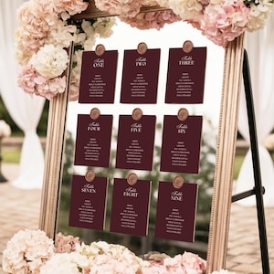Peut inclure: Un miroir doré encadre neuf cartons de table bordeaux pour un mariage. Chaque carton est numéroté et présente des noms. Le miroir est orné de fleurs roses et blanches, créant une présentation élégante.