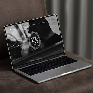 May include: A silver laptop displaying a website with a black and white image and the initials "CD" inside an ornate frame. The laptop is open on a brown surface, with a dark background.