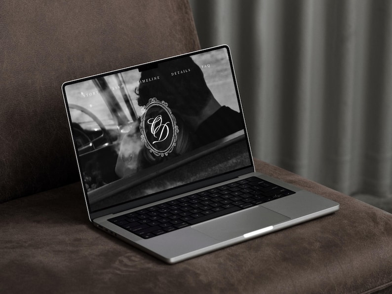 May include: A silver laptop displaying a website with a black and white image and the initials "CD" inside an ornate frame. The laptop is open on a brown surface, with a dark background.