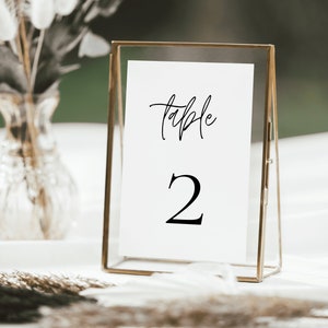 May include: A gold-toned, triangular table number holder with a white card inside. The card has the text "table" written in black cursive script and the number "2" in black font.